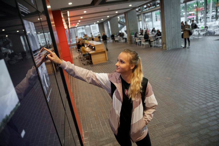 A student uses a touch screen in the campus center’s Moise Y. Safra Welcome Pavilion to look up past Harvard Gazette articles.